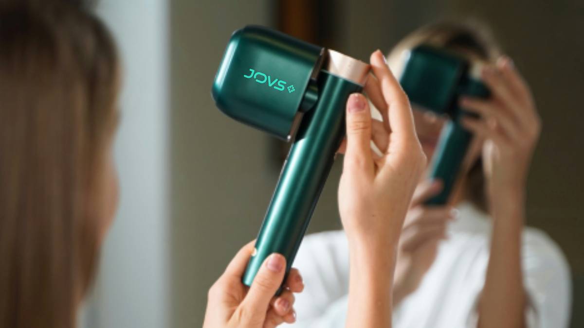 A woman holds the JOVS Venus Pro II Hair Removal Device, with its reflection visible in the mirror.
