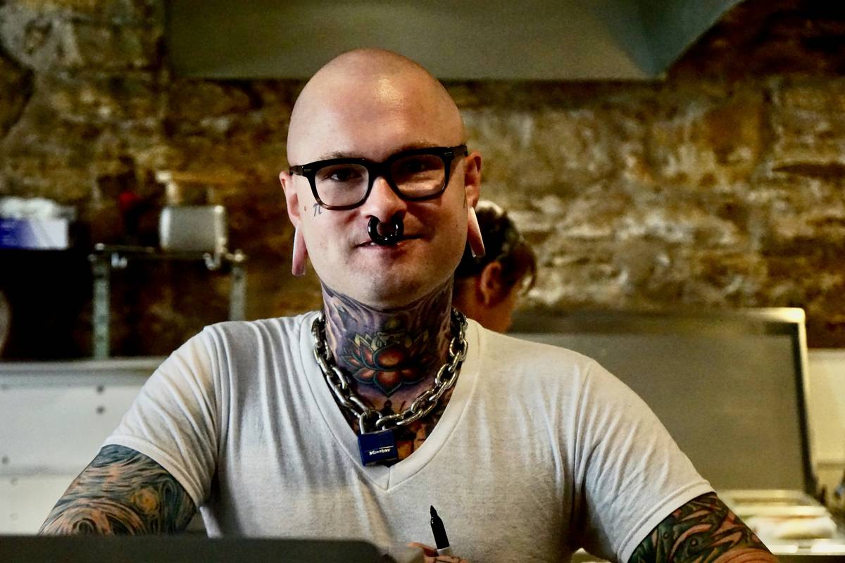 Tattoed bespectacled bald young butcher with nose ring and chains. Looking directly at the camera.